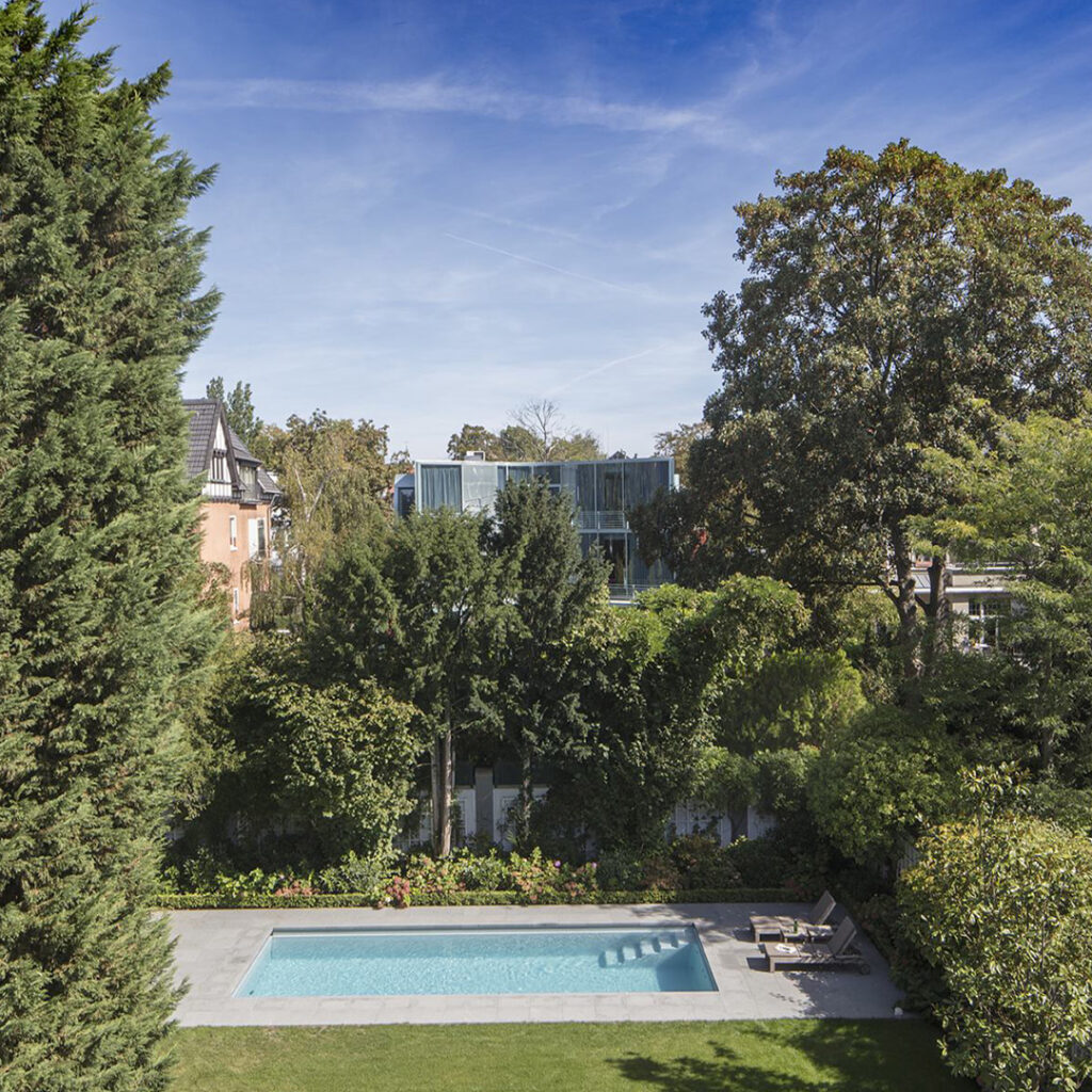 Ein moderner Pool in einem grünen Garten mit Bäumen und blühenden Pflanzen bei sonnigem Wetter.
