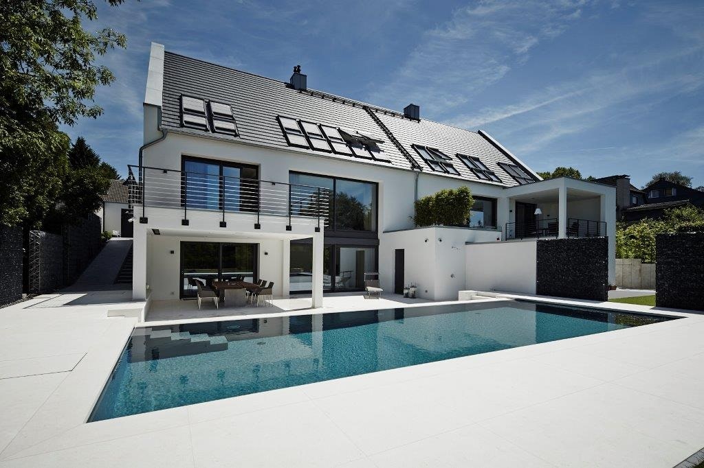 Moderne Villa mit weißer Fassade und großem Pool im Vordergrund, blauer Himmel im Hintergrund.