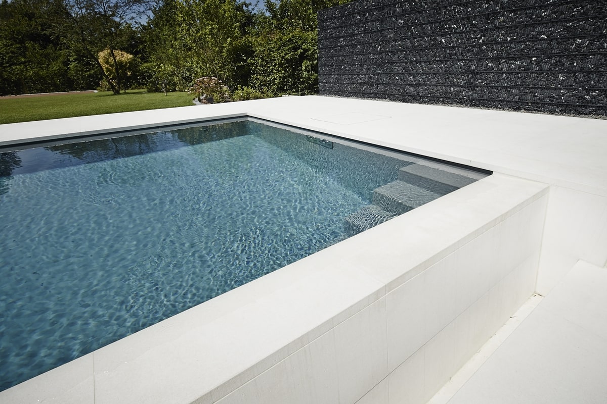 Moderner Swimmingpool mit klarblauem Wasser, umgeben von weißem Beton, mit Garten im Hintergrund.