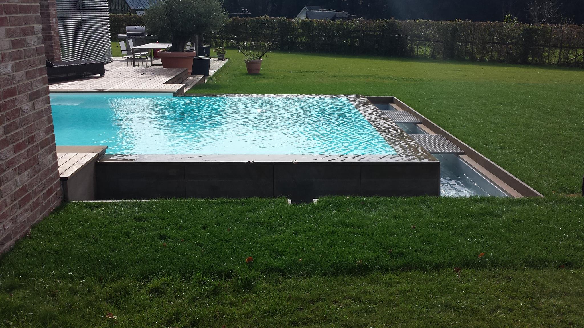 Moderner Pool mit Überlauf, klar blaues Wasser, Holzdeck, grüner Rasen im Hintergrund.