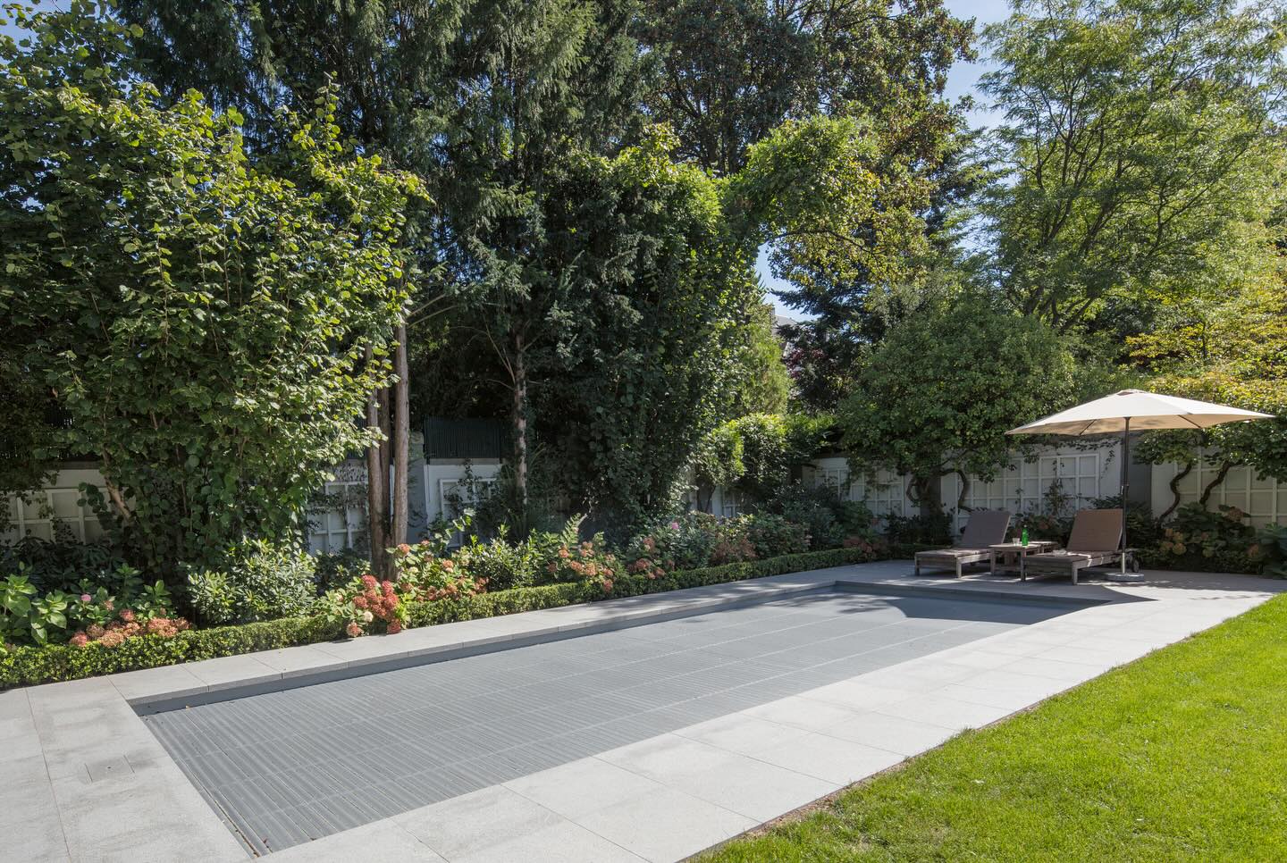 Ein moderner, rechteckiger Pool in einem Garten mit Liegen und einem Sonnenschirm.