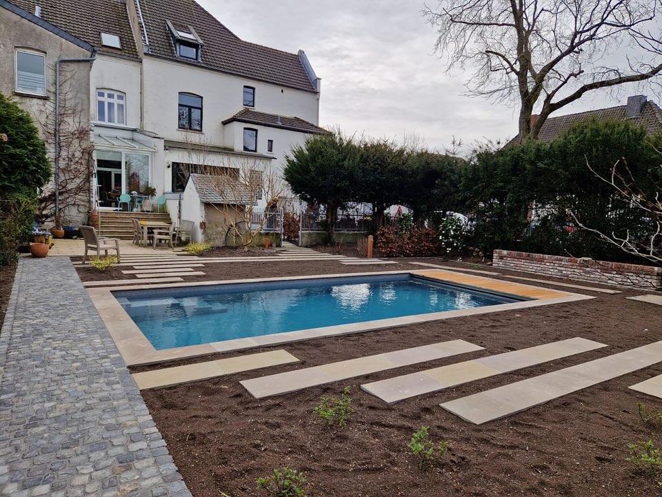 Ein modernes Schwimmbad im Garten eines grauen Hauses, umgeben von Pflanzen.