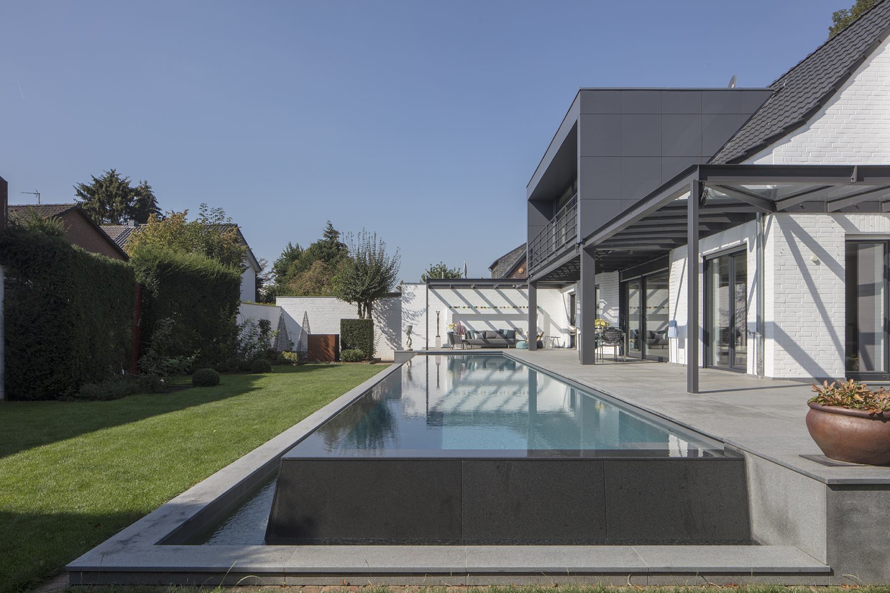 Moderne Villa mit Pool und Garten, klare Linien, graue und weiße Fassaden, strahlend blauer Himmel.