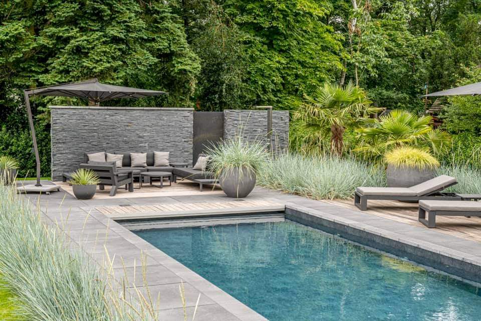 Moderner Garten mit Pool, grauen Liegestühlen, Pflanzen und einer Steinmauer im Hintergrund.