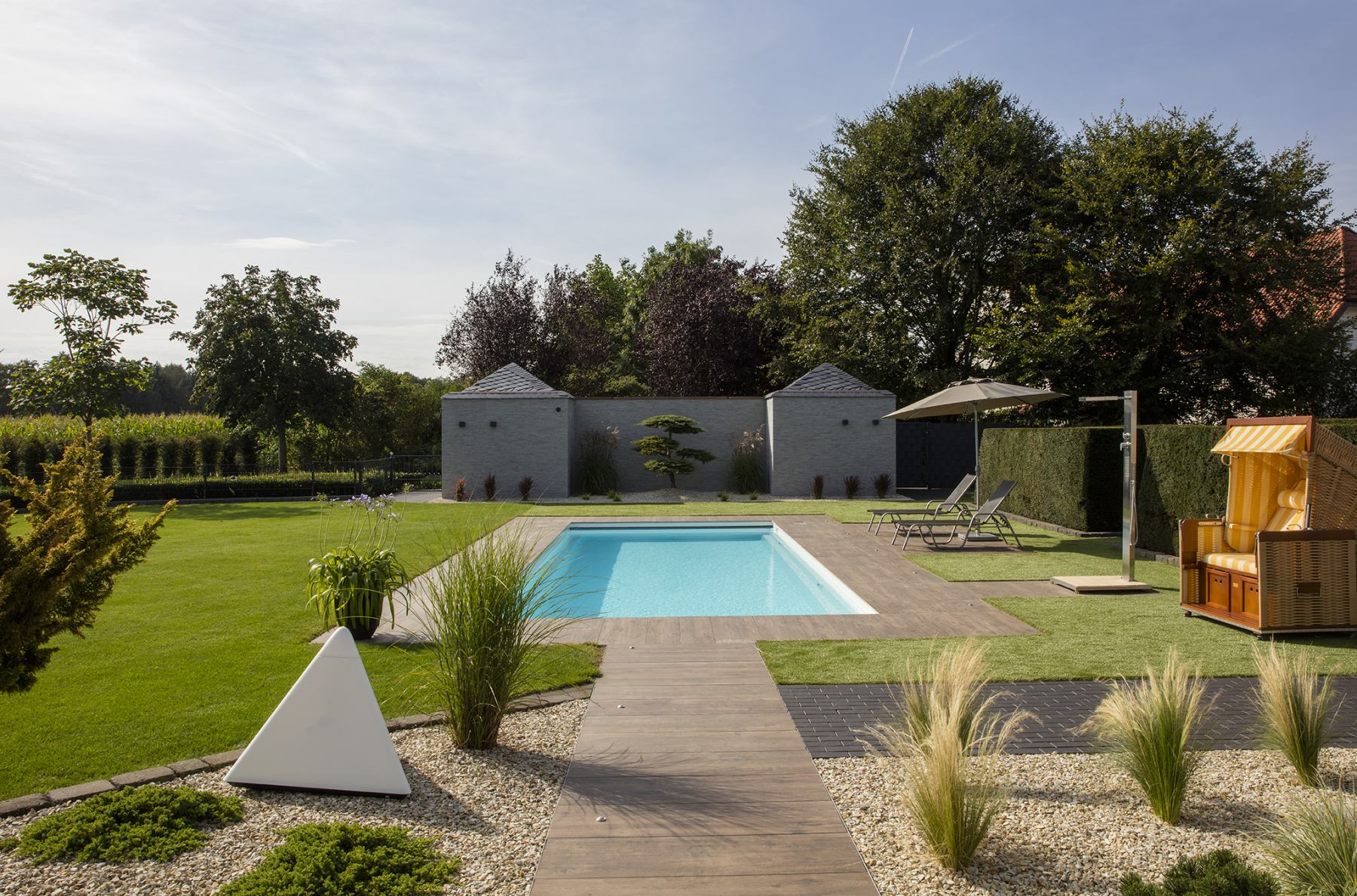 Ein moderner Garten mit Pool, grüner Rasen, Kieselwege und Sonnenliegen bei hellem Tageslicht.