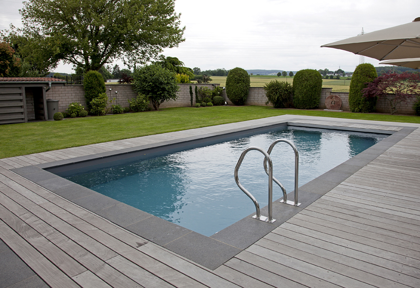 Rechteckiger Pool im Garten, umgeben von Holzterrasse, grüner Rasen und Hecken im Hintergrund.