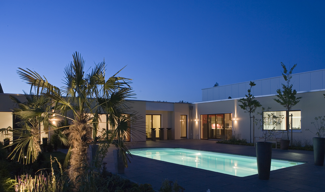Beleuchtetes modernes Haus mit Pool und Palmen, Dämmerungshimmel im Hintergrund.