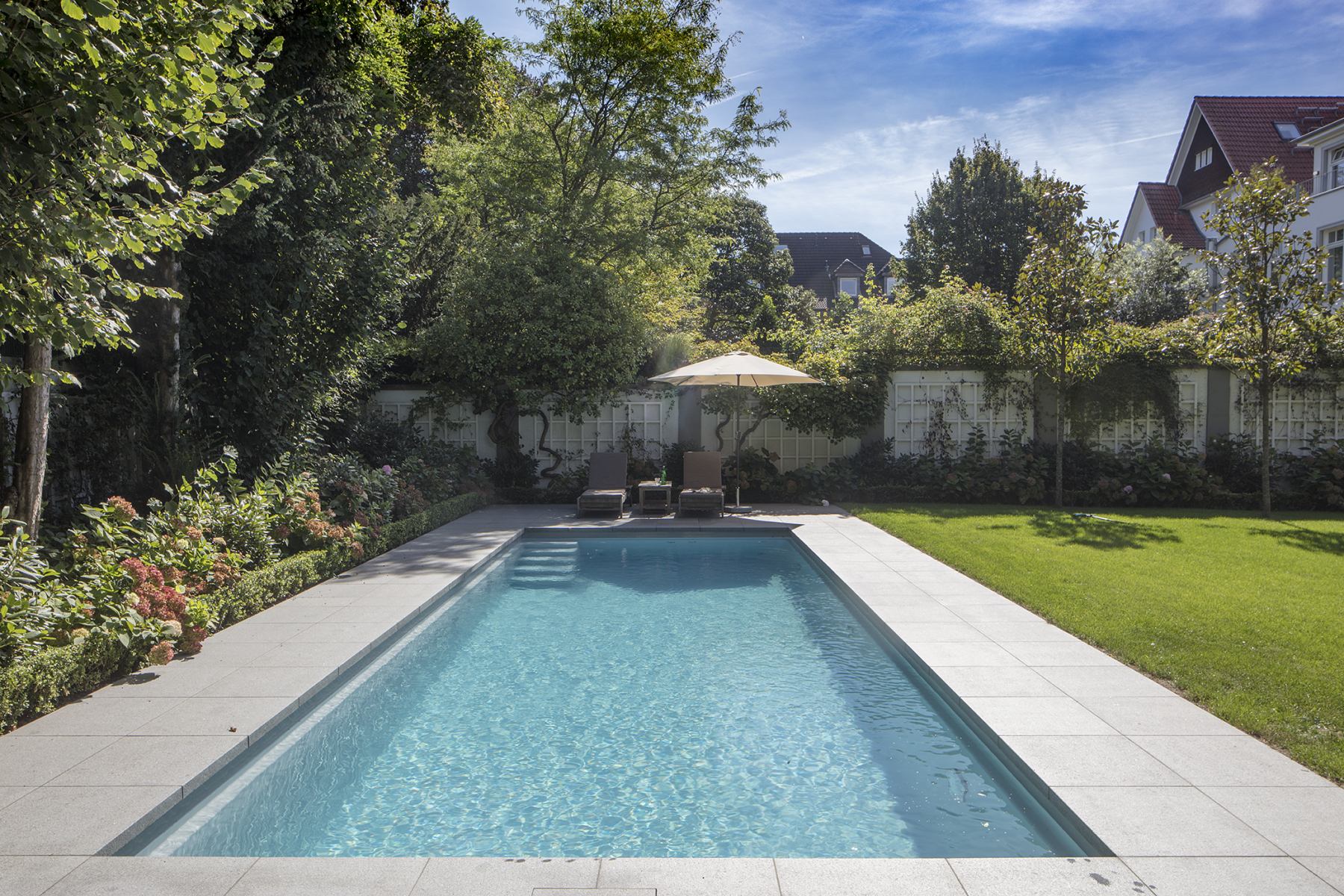 Ein Garten mit Swimmingpool, grünen Bäumen und Rasen, sonniges Wetter, zwei Liegestühle mit Schirm.