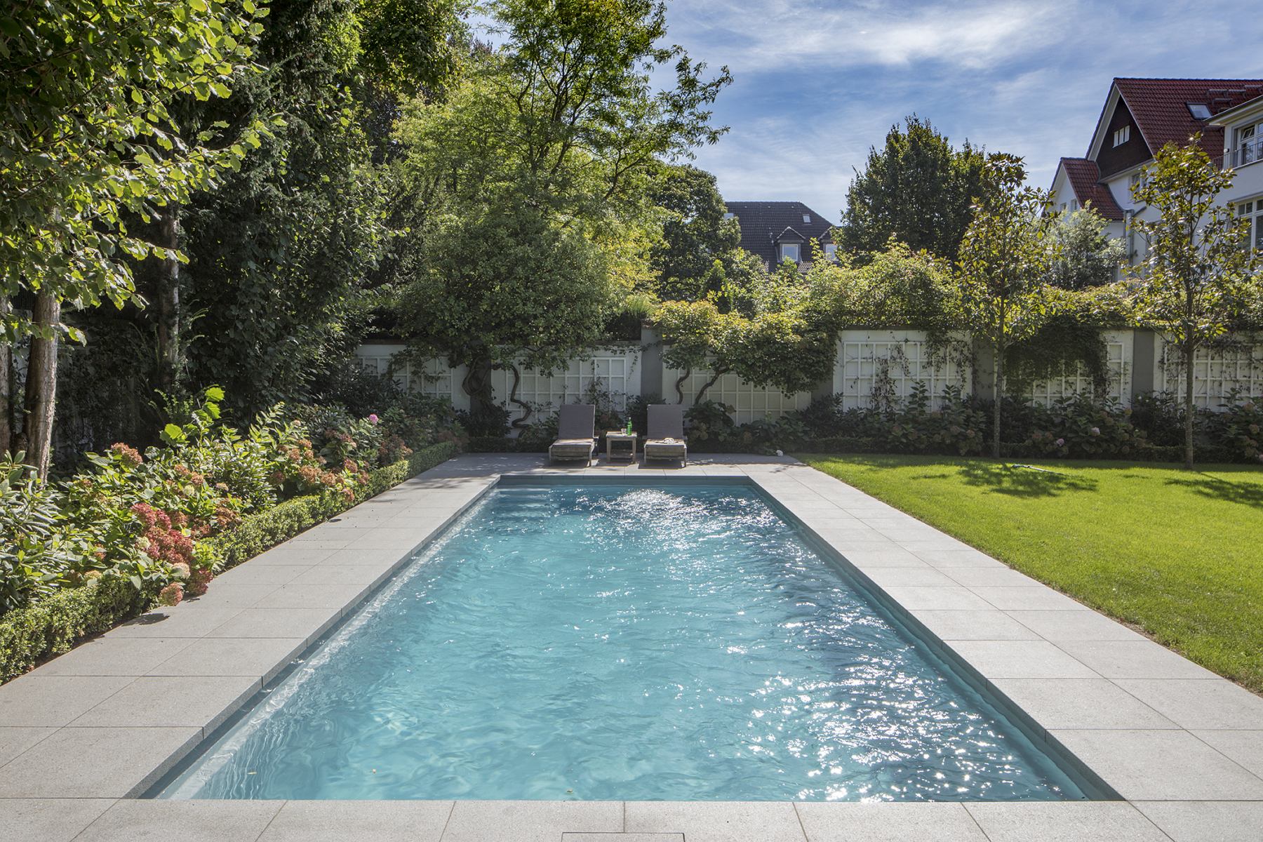 Rechteckiger Pool im Garten, umgeben von Bäumen und Blumen, mit zwei Liegestühlen im Hintergrund.