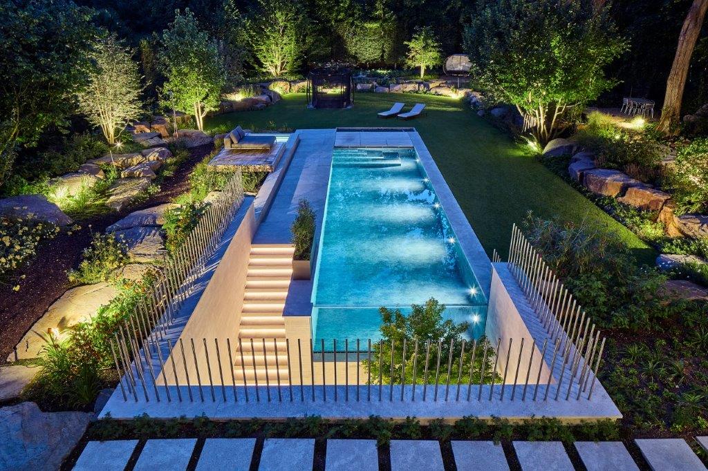 Beleuchteter moderner Pool in einem Garten mit Liegestühlen und vielen Bäumen im Hintergrund.