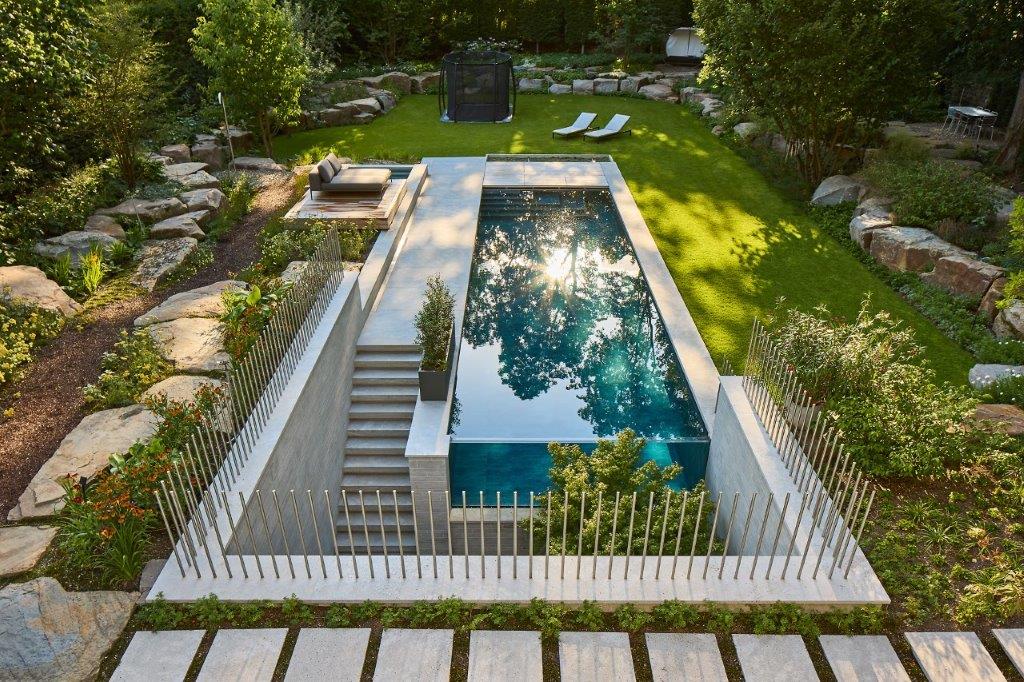 Ein moderner Garten mit rechteckigem Pool, umgeben von Pflanzen und Rasen. Sonnenreflexionen im Wasser.
