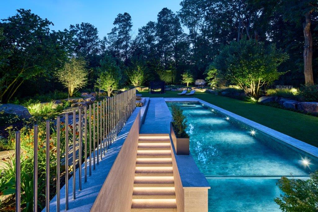 Beleuchteter moderner Garten mit Pool, Treppe und Bäumen im Abendlicht.