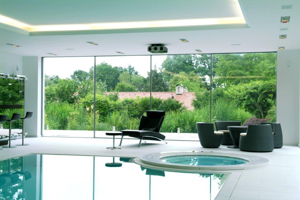 Innenpool mit großen Fenstern, Naturblick, schwarze Stühle, Liege, modernes Ambiente.