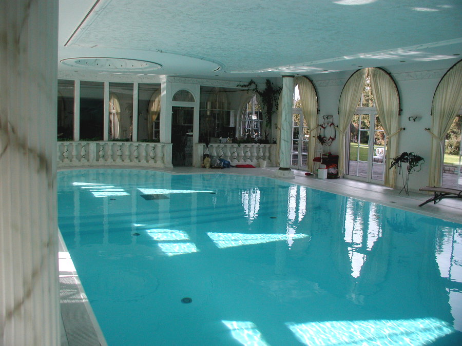 Innenraum mit großem, hellem Pool, weißen Säulen und eleganten Vorhängen. Keine Personen zu sehen.