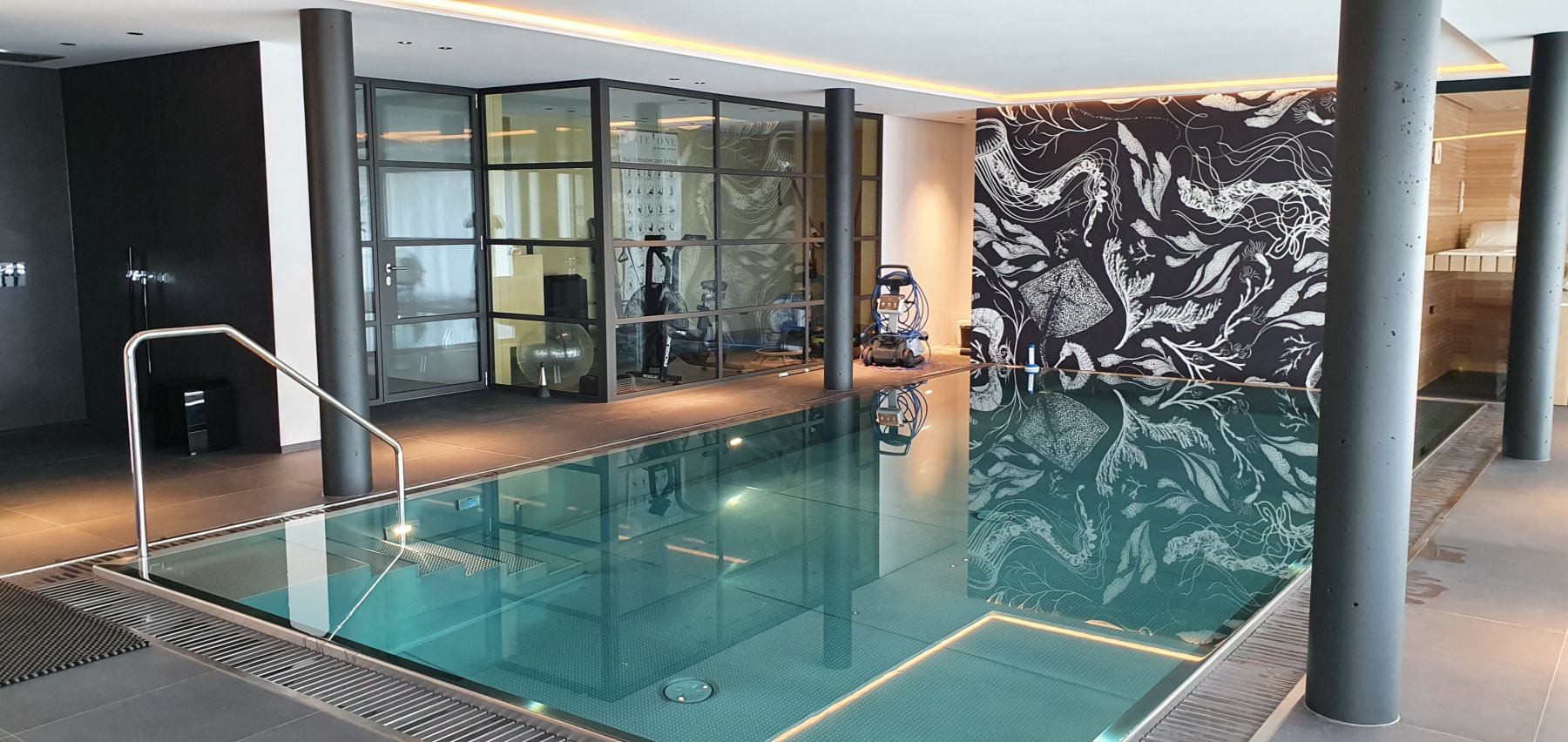 Ein Hallenbad mit klarem Wasser, stilvolle Wandgrafik, moderner Fitnessraum im Hintergrund.