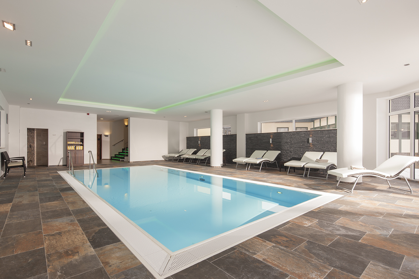 Innenpoolbereich mit modernem Design, graue Fliesen, Liegen um den Pool, helles Beleuchtungskonzept.