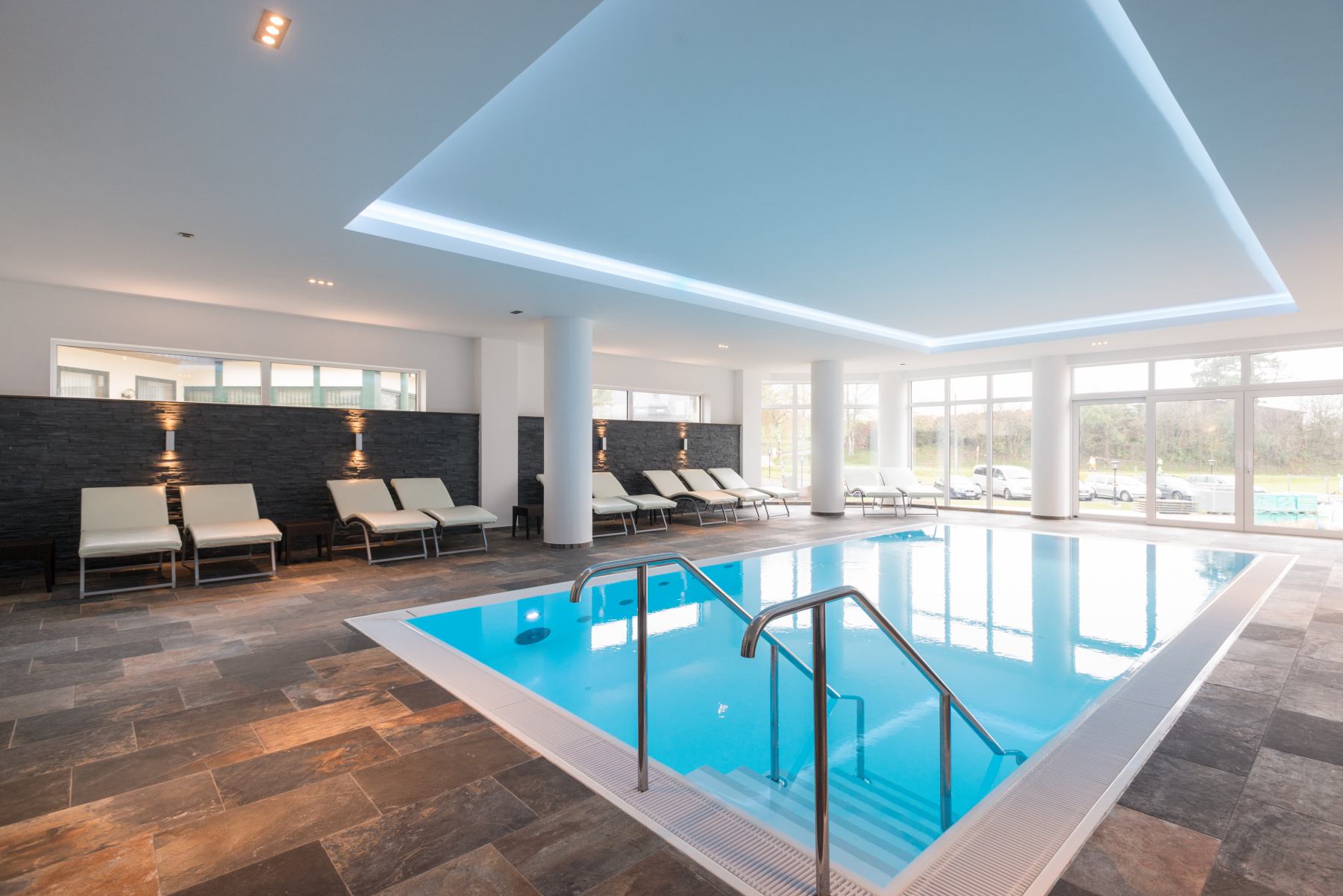 Innenpool mit Liegestühlen, modern gestaltet, großen Fenstern und Deckeneinbauleuchten.