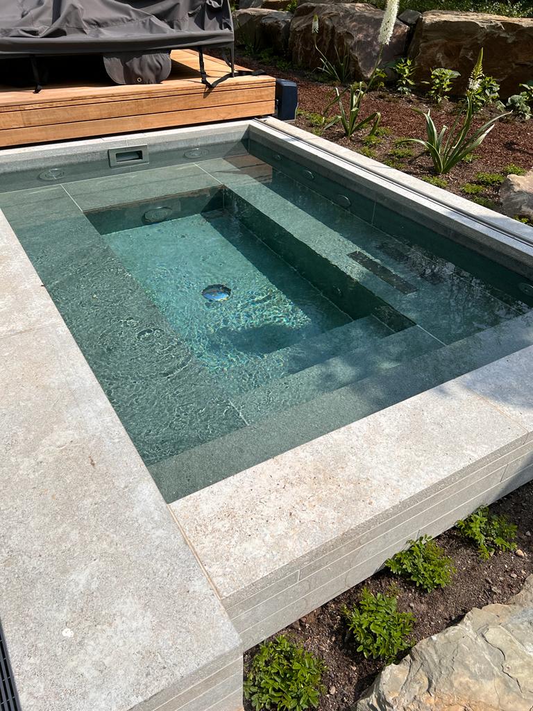 Kleiner, rechteckiger Pool im Freien, umgeben von Steinen und Pflanzen, mit klarem Wasser.