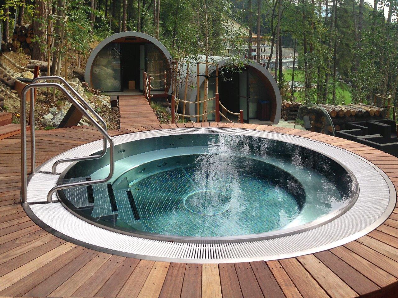 Ein runder Whirlpool im Wald, umgeben von Holzterrasse, mit futuristischen Gebäuden im Hintergrund.