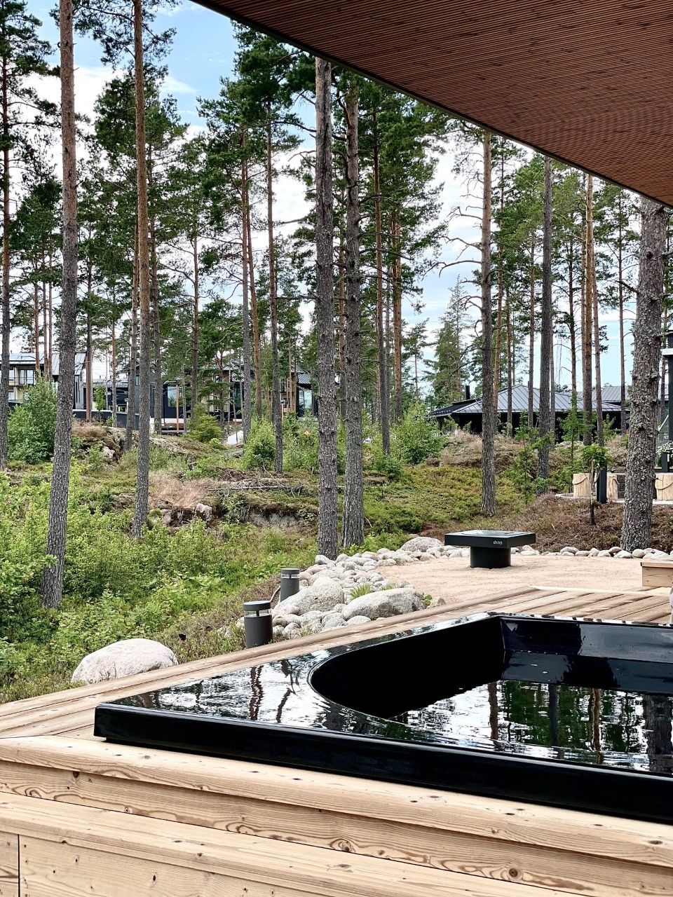 Holzveranda mit schwarzem Whirlpool im Wald, umgeben von hohen Bäumen und grüner Vegetation.