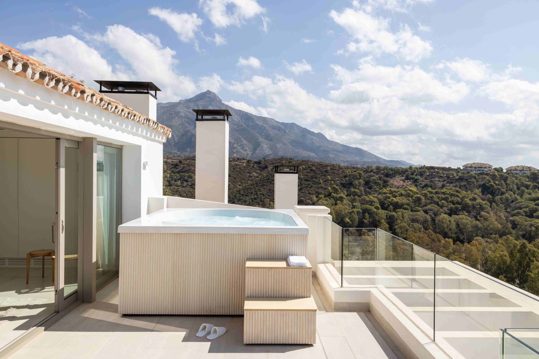 Dachterrasse mit Whirlpool, Berge im Hintergrund, sonnig, moderne Glasgeländer, weiß-beige Akzente.