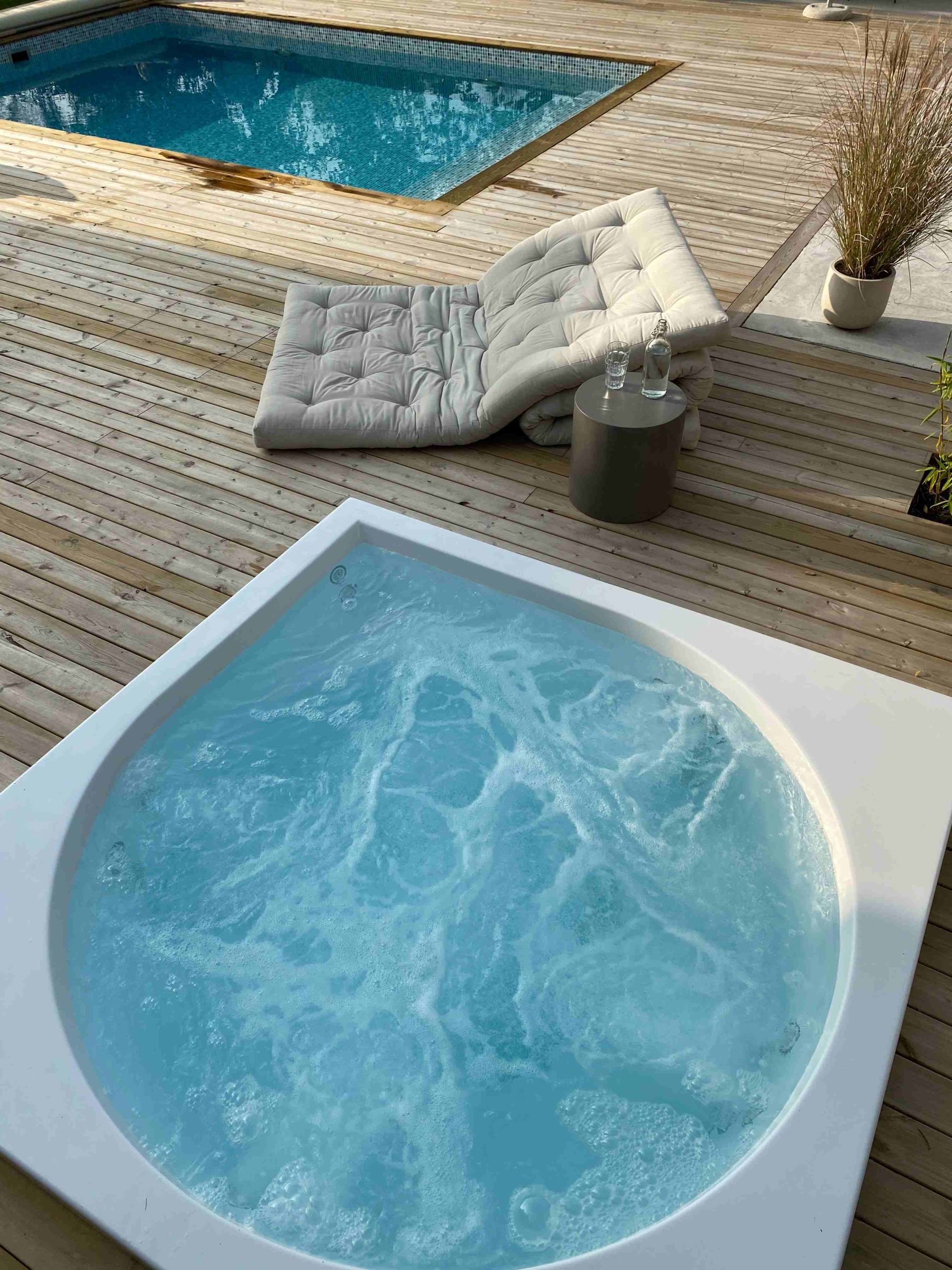 Whirlpool auf Holzterrasse, daneben zwei Kissen und ein Tisch mit Gläsern, Pool im Hintergrund.