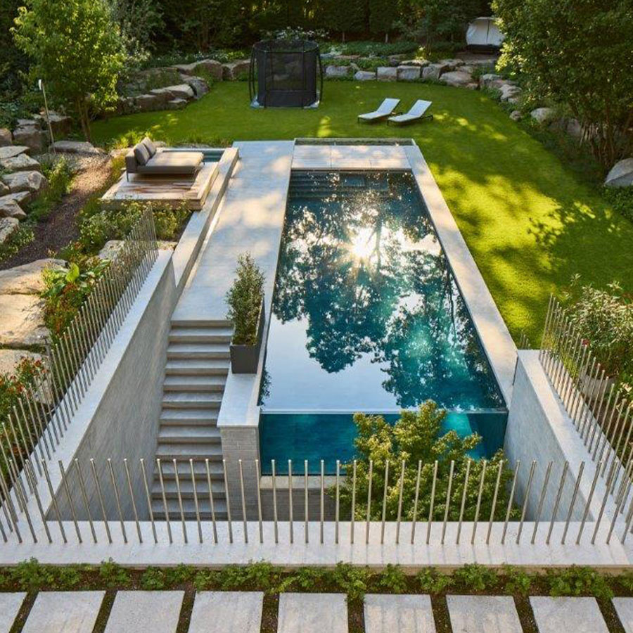 Rechteckiger Pool mit Reflektionen, umgeben von Garten, Liegestühlen und Zaun im Sonnenlicht.
