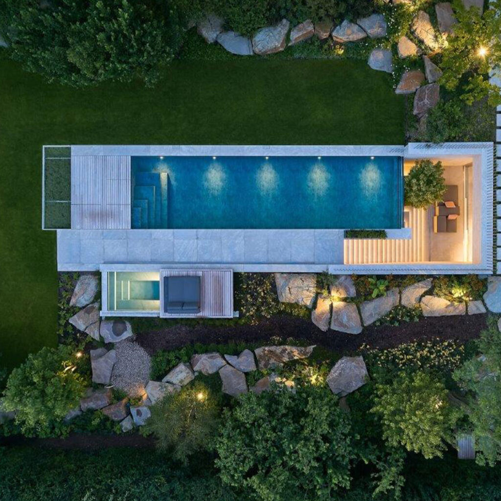 Luftaufnahme eines rechteckigen Pools mit umgebender Gartenlandschaft und Steinen, abends beleuchtet.