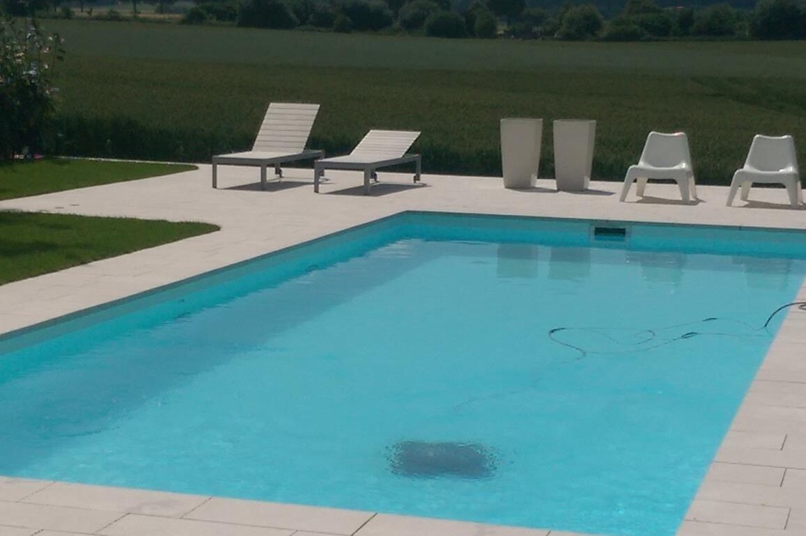 Ein leerer Swimmingpool mit blauem Wasser, umgeben von weißen Liegestühlen auf Steinfliesen.