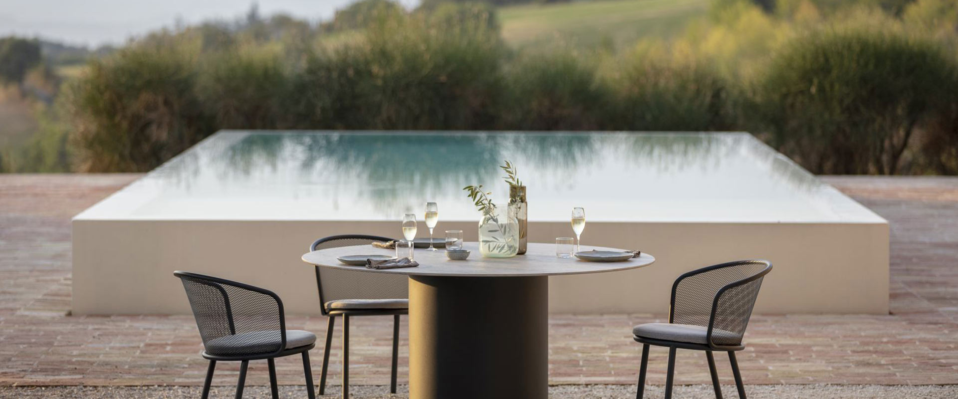 Runder Tisch mit Weingläsern, drei moderne Stühle, Pool und Landschaft im Hintergrund.