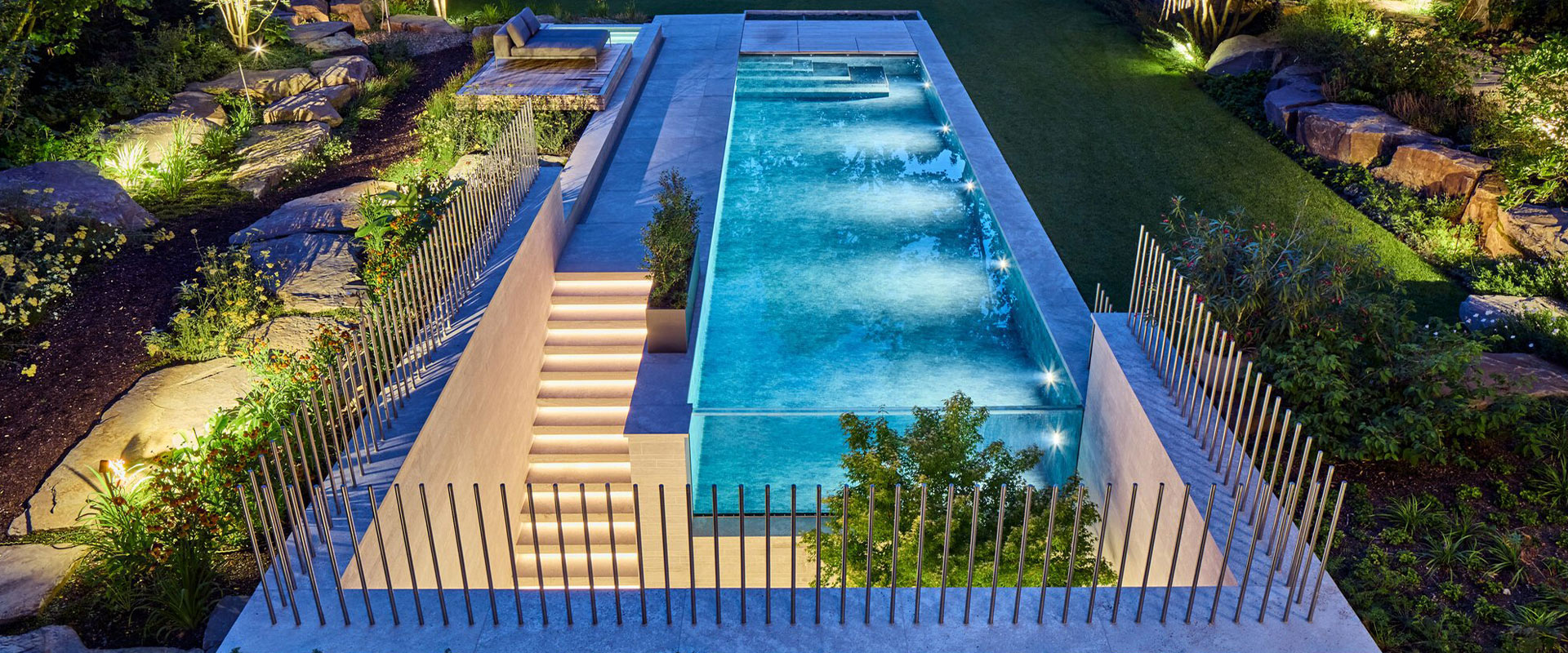Beleuchteter Pool im Garten, umgeben von Pflanzen; moderne Architektur, abends.