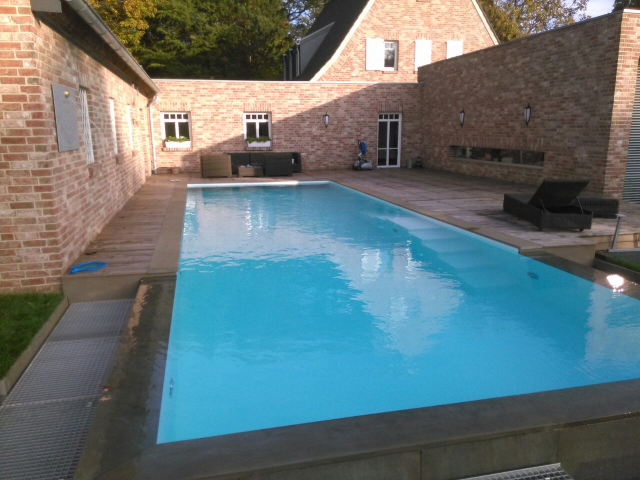 Backsteinhaus mit großem rechteckigem Pool, umgeben von Liegestühlen, bei sonnigem Wetter.