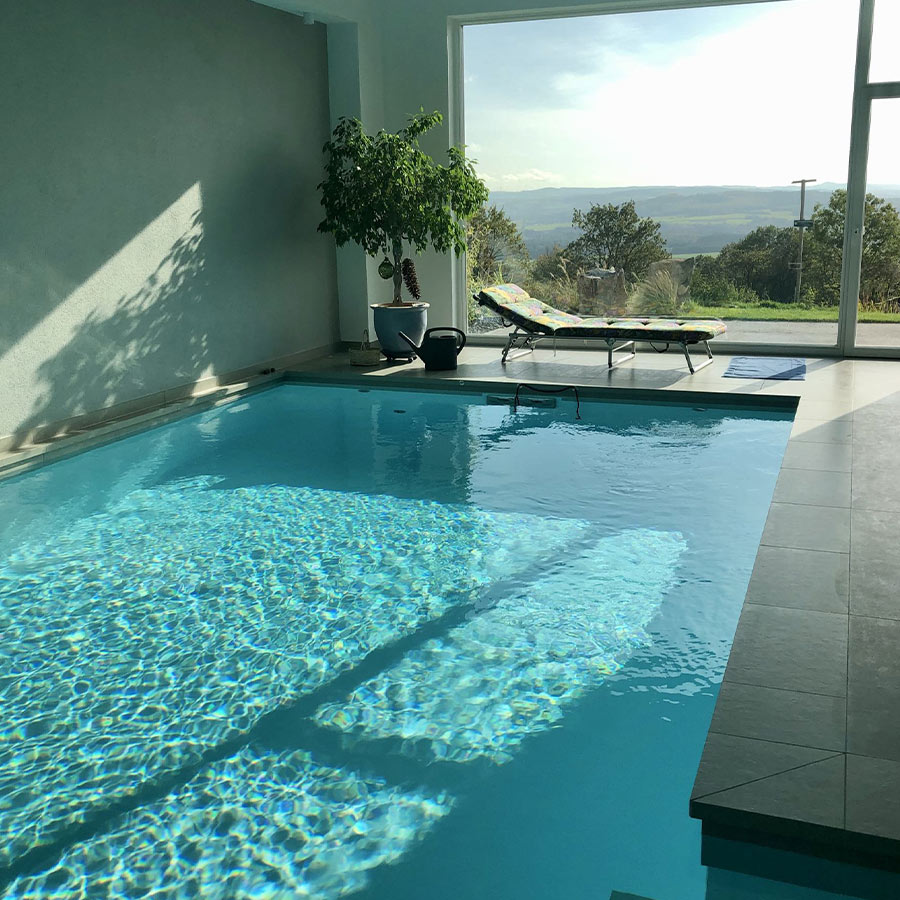 Innenpool mit klarem Wasser, helle Wände, grüne Pflanze, Liegestuhl, großes Fenster mit Landschaft.