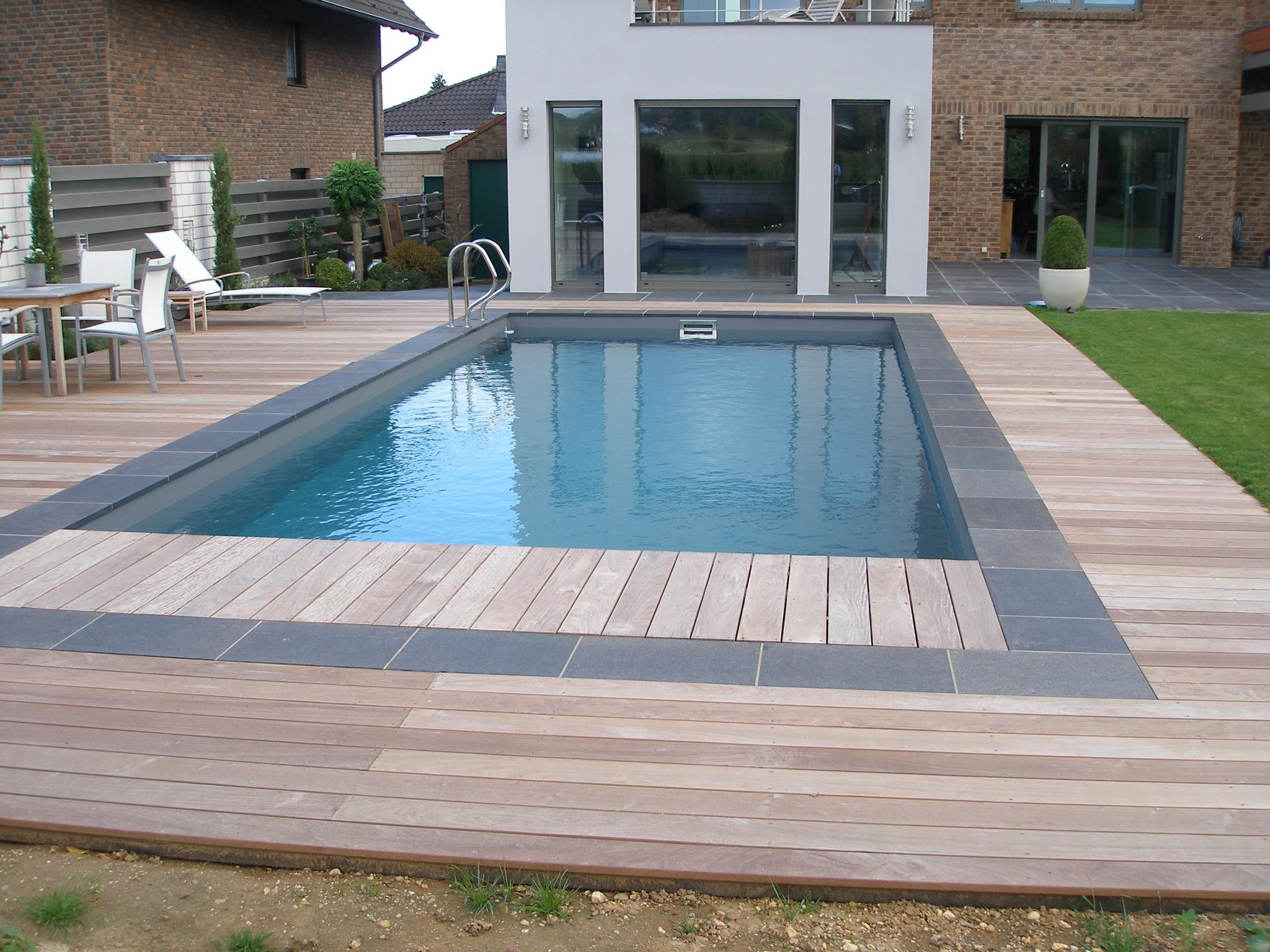Moderner Pool mit Holzterrasse, neben einem Haus mit Steinwänden und großen Fenstern.