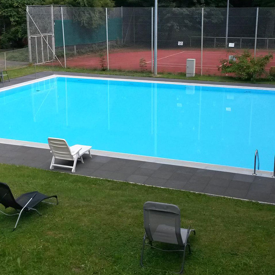 Ein leerer Außenpool mit blauen Wasser, umgeben von wenigen Liegestühlen und einem Tennisplatz.