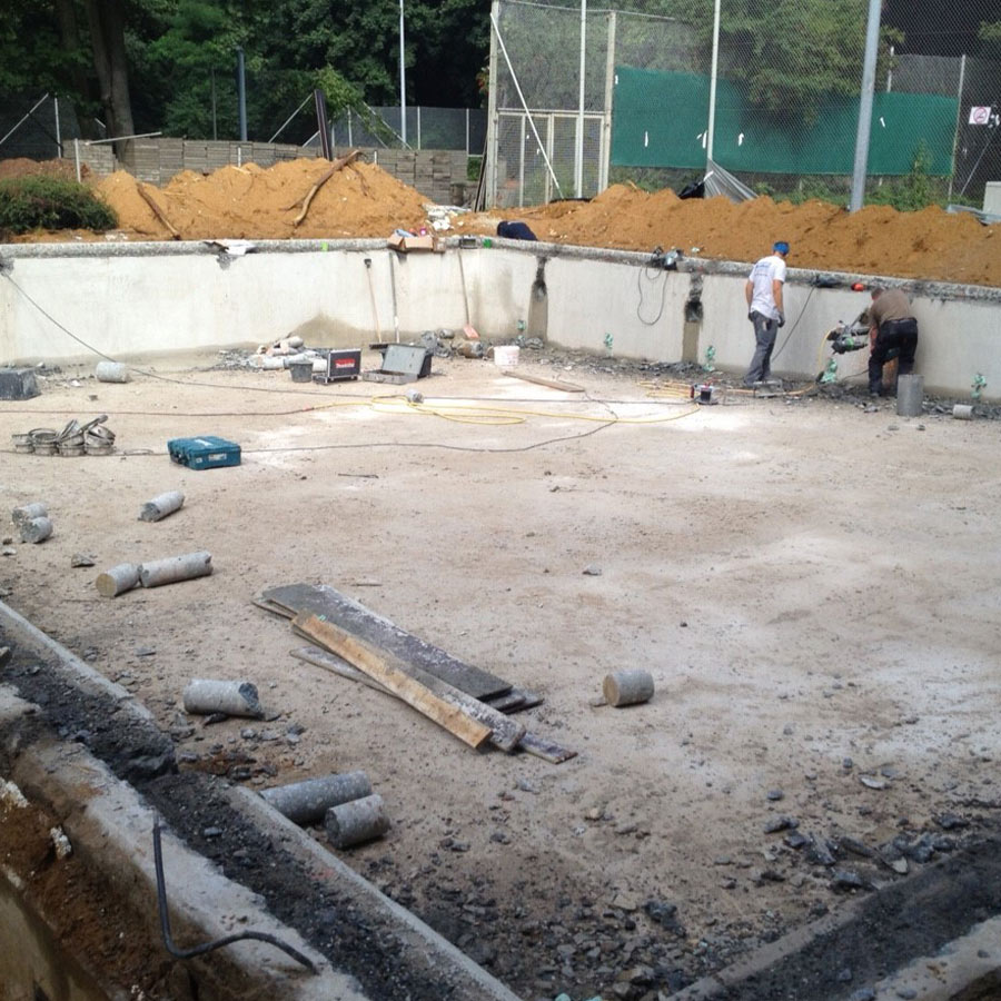 Zwei Arbeiter renovieren ein leeres Schwimmbecken, umgeben von Baumaterialien, im Freien.