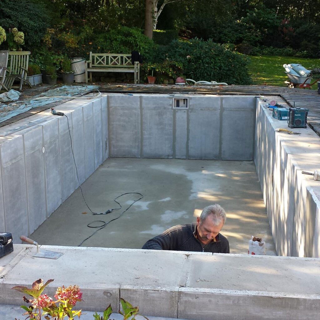 Eine Person mit ernstem Ausdruck arbeitet an einem leeren grauen Pool, umgeben von Gartenmöbeln.