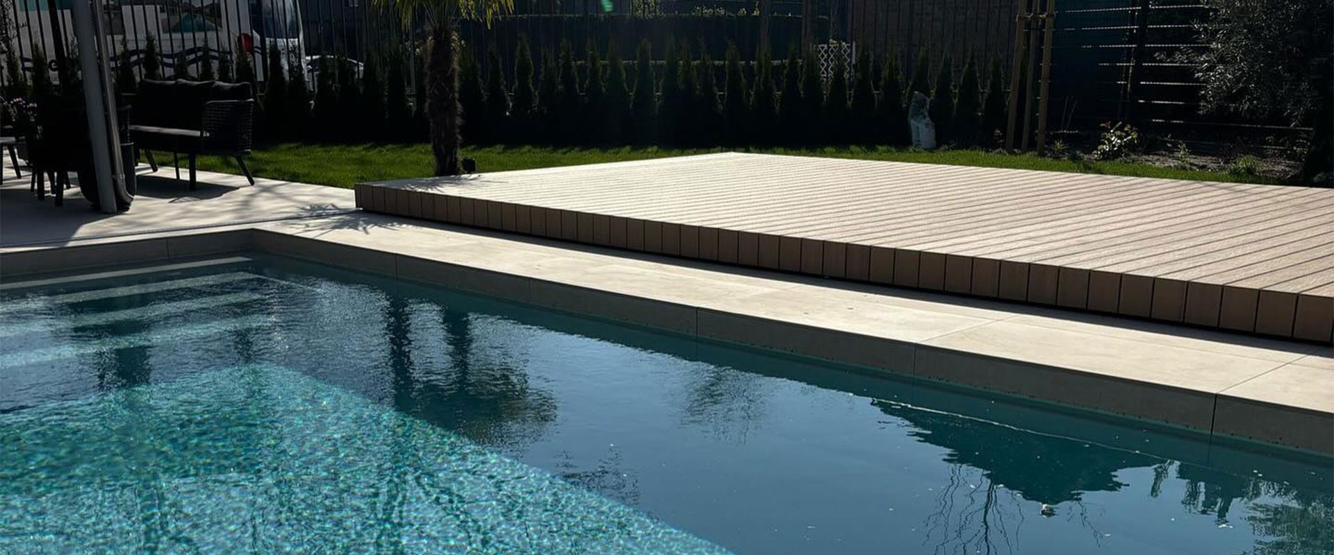 Ein Pool im Garten mit Holzboden und dunklen Möbeln bei sonnigem Wetter.
