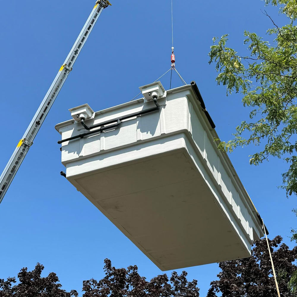 Ein großer weißer Container schwebt am Kranhaken vor blauem Himmel und grünen Bäumen.