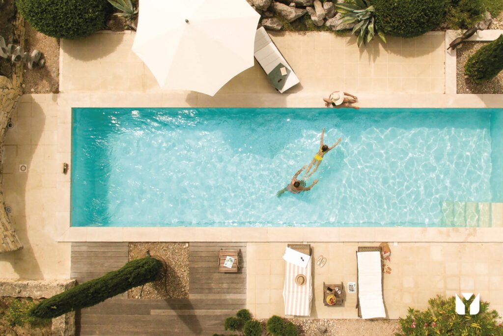 Eine Person schwimmt im blauen Pool unter Sonnenschirm auf hellem Boden.